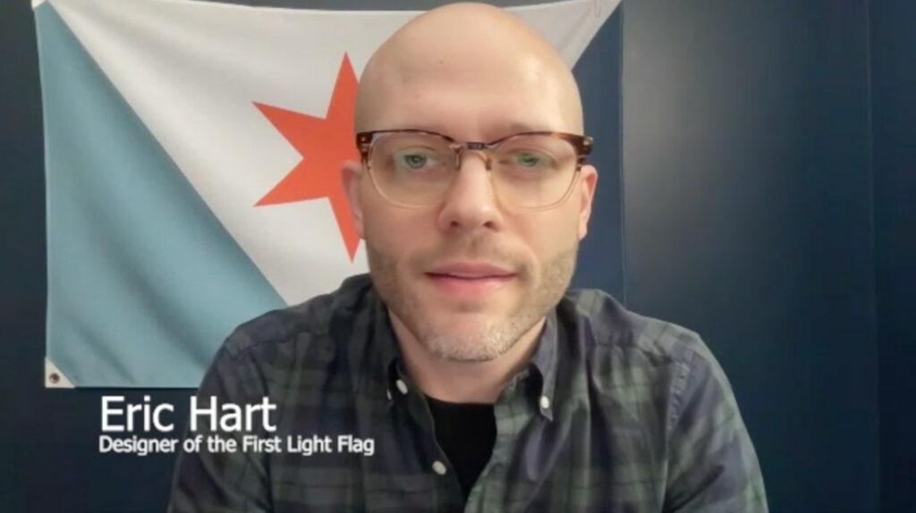 Video interview still of Eric Hart, wearing glasses and a plaid shirt, with the Syracuse city flag hanging behind him.
