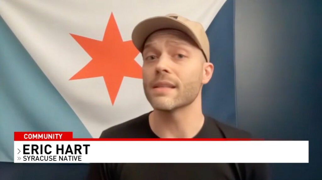 CNY Central news interview still of Eric Hart wearing a tan cap, speaking on camera with the Syracuse city flag in the background.