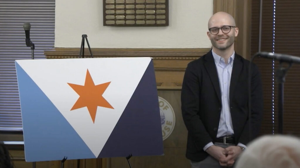 Eric Hart standing beside a printed poster of the Syracuse city flag design during a presentation at Syracuse City Hall