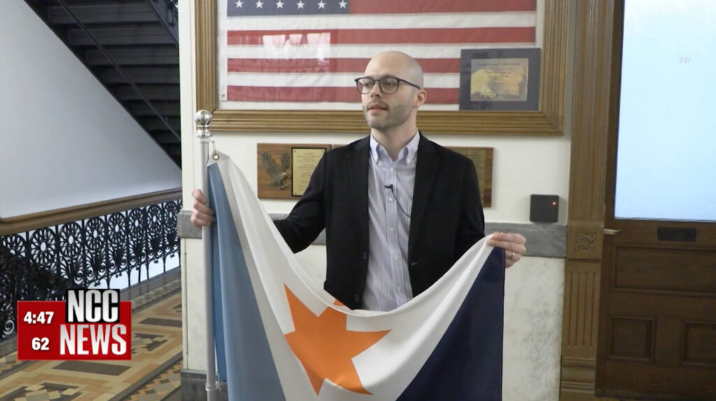 NCC News interview still of Eric Hart in a blazer, holding up and unfurling the Syracuse city flag.