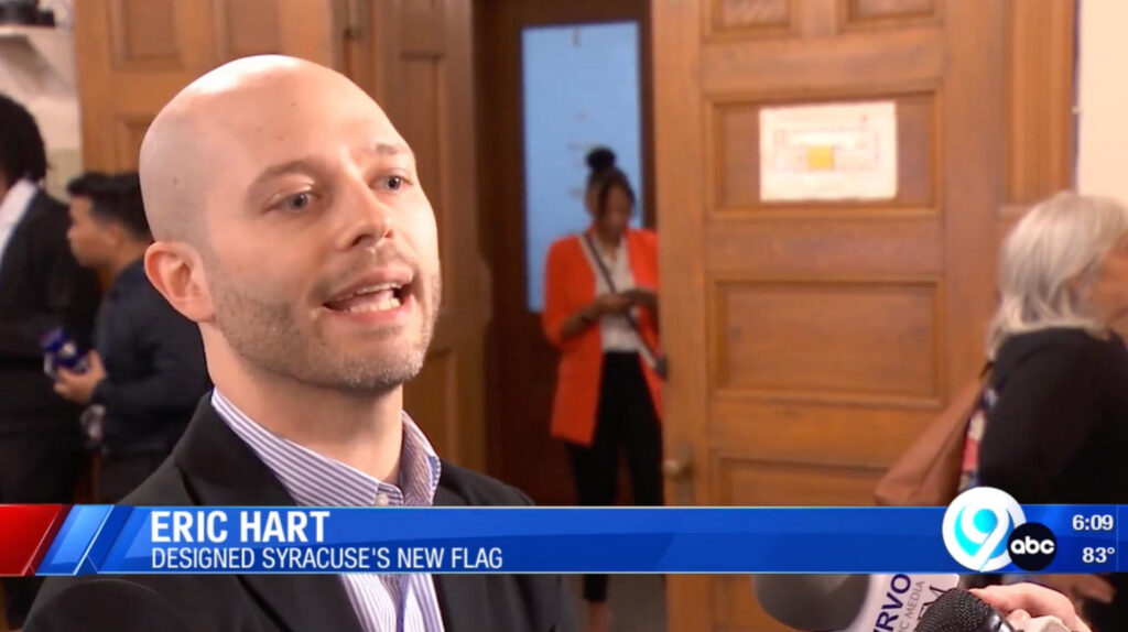 News Channel 9 interview still of Eric Hart speaking to reporters about the Syracuse city flag indoors, being recorded with microphones.