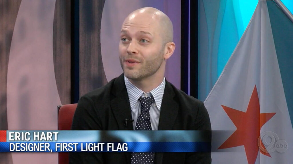 News Channel 9 "Newsmakers" studio interview still of Eric Hart in a suit and tie, seated in front of a large Syracuse city flag backdrop.
