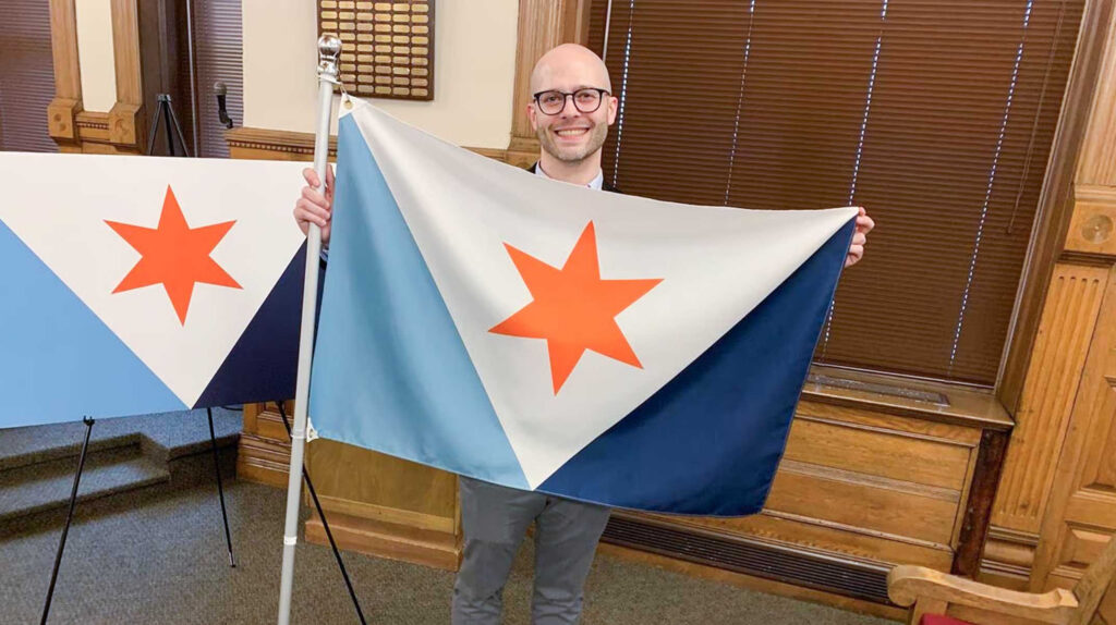 Eric Hart smiling and holding up the Syracuse city flag on a pole indoors