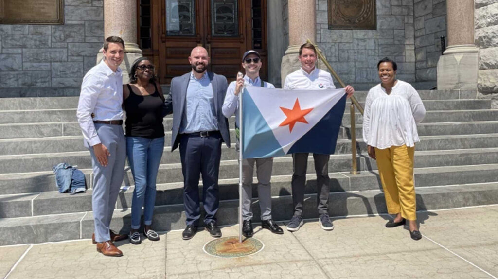 Group of city officials smiling on the steps of Syracuse City Hall, gathered around the Syracuse city flag