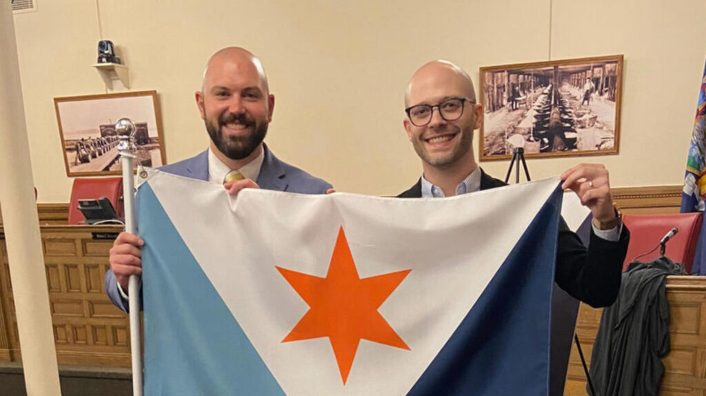 Eric Hart and Andrew Frasier smiling indoors as they hold up the Syracuse city flag together