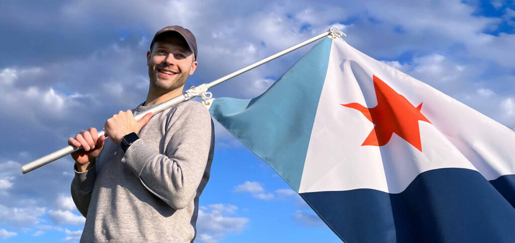 Eric Hart, designer of the Syracuse city flag, holding the flag on a pole against a blue sky.