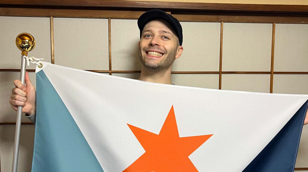 Eric Hart smiling and holding up the Syracuse city flag on a pole indoors, peeking over the top edge of the flag.