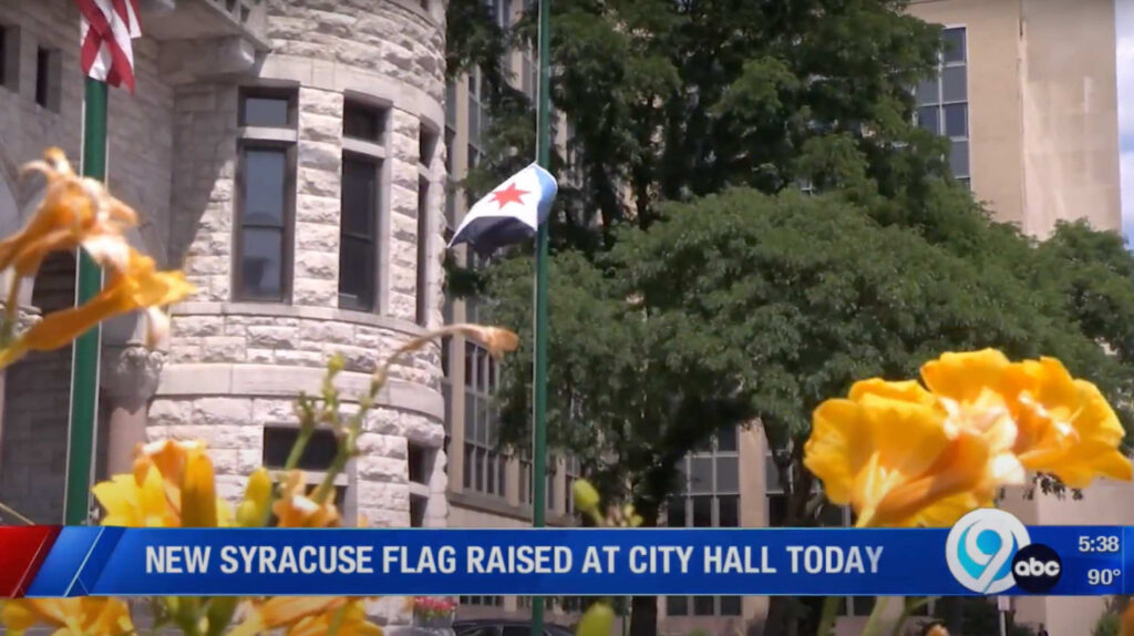 News Channel 9 broadcast still of the Syracuse city flag flying at City Hall, seen through yellow flowers in the foreground.