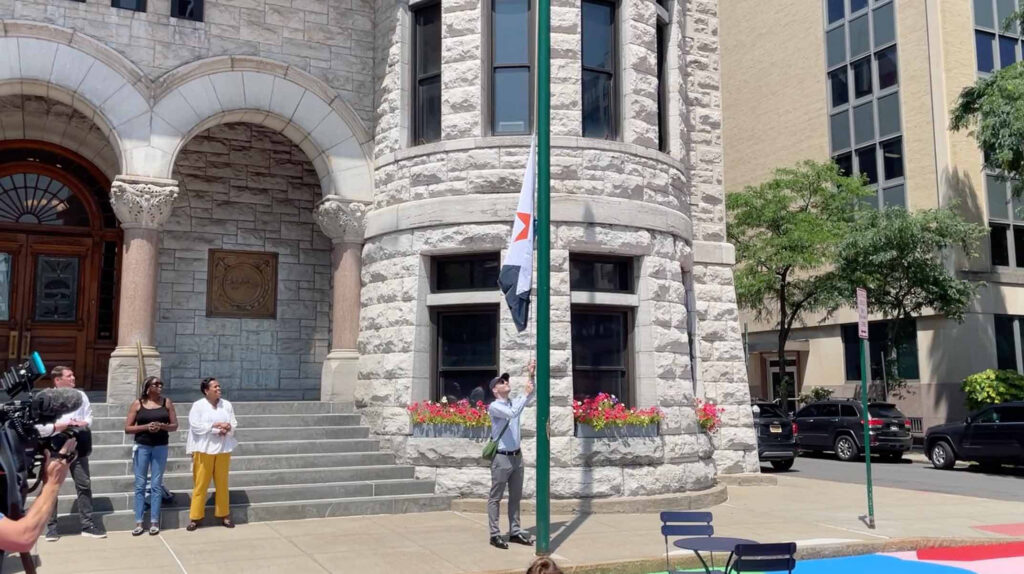 Outdoor photo of a flag-raising ceremony at Syracuse City Hall, showing Eric Hart raising the Syracuse city flag up a pole as a small crowd, camera operator, and city officials look on.