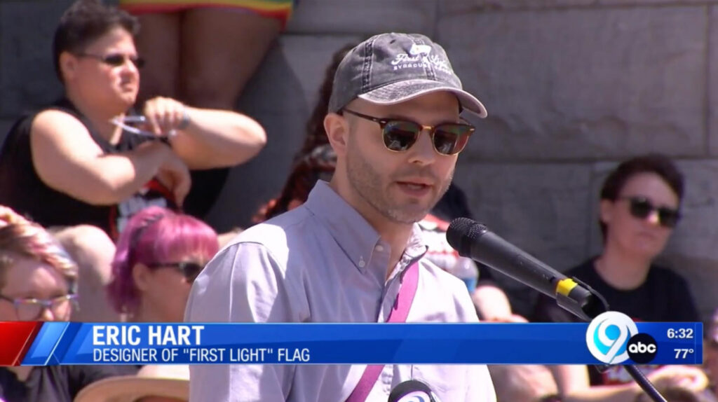 News Channel 9 outdoor broadcast still of Eric Hart, wearing a First Light Syracuse Goods cap and sunglasses, speaking at a microphone to a crowd.