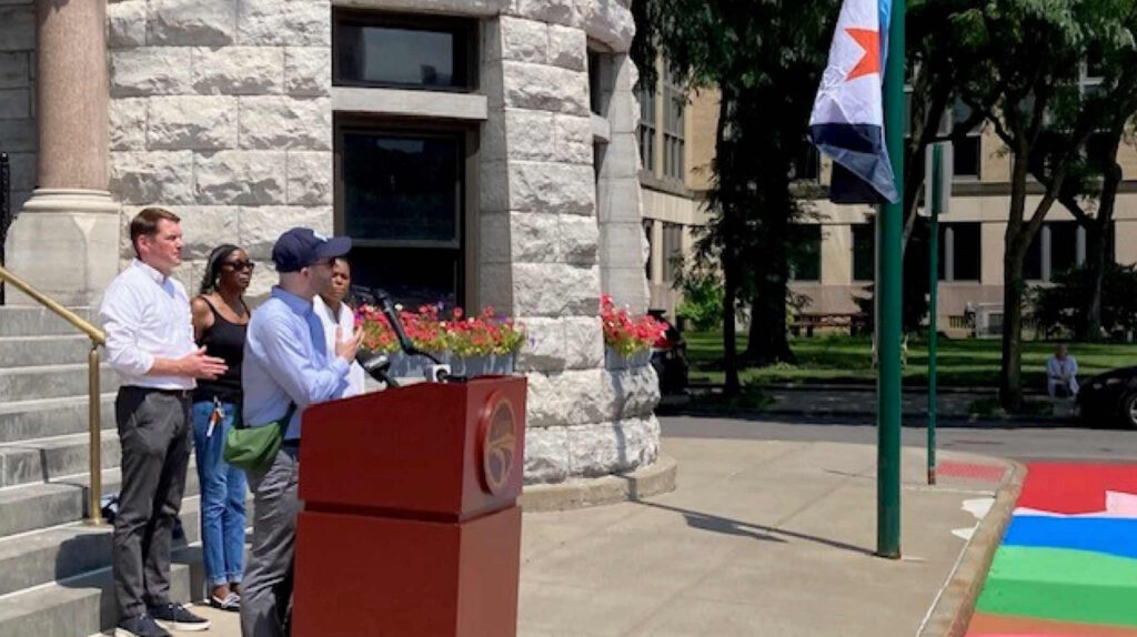 Eric Hart and city officials speaking at a podium outside Syracuse City Hall during the flag-raising ceremony, with the Syracuse city flag flying on a pole nearby.